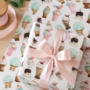 May include: A wrapped gift with a light pink satin bow. The wrapping paper is white and covered in colorful illustrations of smiling ice cream cones, sundaes, and milkshakes. A spool of pink ribbon is visible in the background.