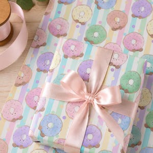 May include: A wrapped gift with a pink satin bow sits on a sheet of wrapping paper. The paper features a repeating pattern of smiling donuts in pastel colors on a striped background. A spool of pink ribbon and greenery are in the background.
