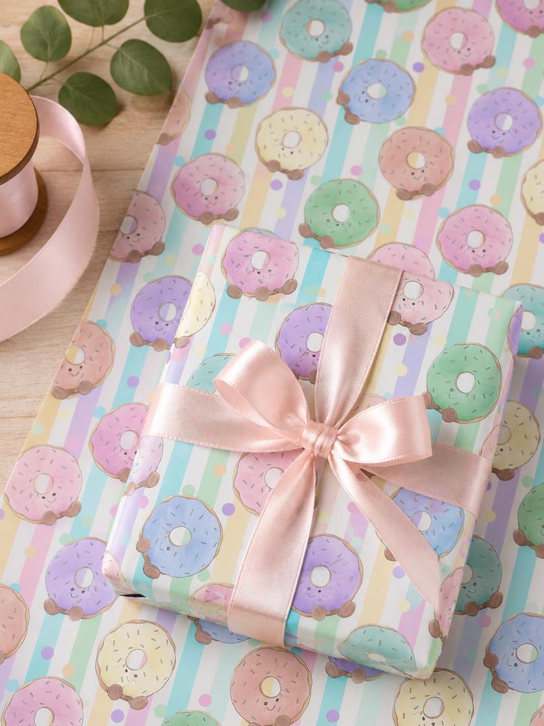 May include: A wrapped gift with a pink satin bow sits on a sheet of wrapping paper. The paper features a repeating pattern of smiling donuts in pastel colors on a striped background. A spool of pink ribbon and greenery are in the background.