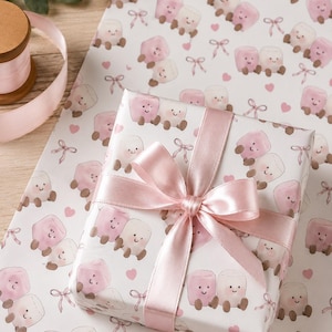 May include: A wrapped gift with a pink satin ribbon and bow sits on a sheet of matching wrapping paper. The paper features a repeating pattern of smiling marshmallow characters in pink and white, with small hearts and bows.