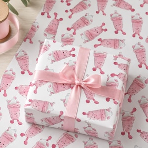 May include: A wrapped gift with a pink satin ribbon and bow, placed on a sheet of matching wrapping paper. The wrapping paper features a repeating pattern of pink milkshake characters with smiling faces on a white background.