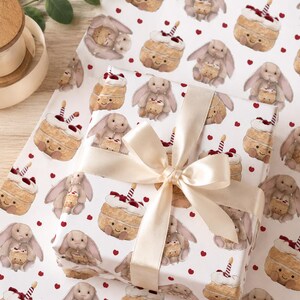 May include: White wrapping paper with a repeating pattern of cartoon bunnies holding cakes and red hearts. A wrapped gift with a cream-colored bow is in the center. The paper is ideal for birthdays or baby showers.