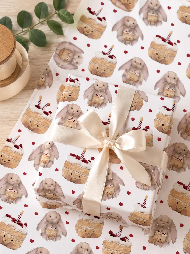 May include: White wrapping paper with a repeating pattern of cartoon bunnies holding cakes and red hearts. A wrapped gift with a cream-colored bow is in the center. The paper is ideal for birthdays or baby showers.