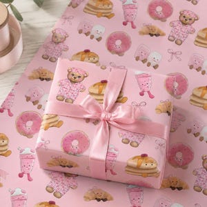 May include: A pink gift box wrapped in patterned paper featuring illustrations of donuts, pancakes, teddy bears, and milkshakes. The box is tied with a pink satin ribbon and bow. The background is a matching pink wrapping paper.