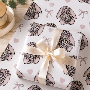 May include: White gift wrap featuring a repeating pattern of leopard print bunny rabbits, pink hearts, and bows. A wrapped gift is tied with a cream-colored satin ribbon. The overall theme is Easter or baby shower.
