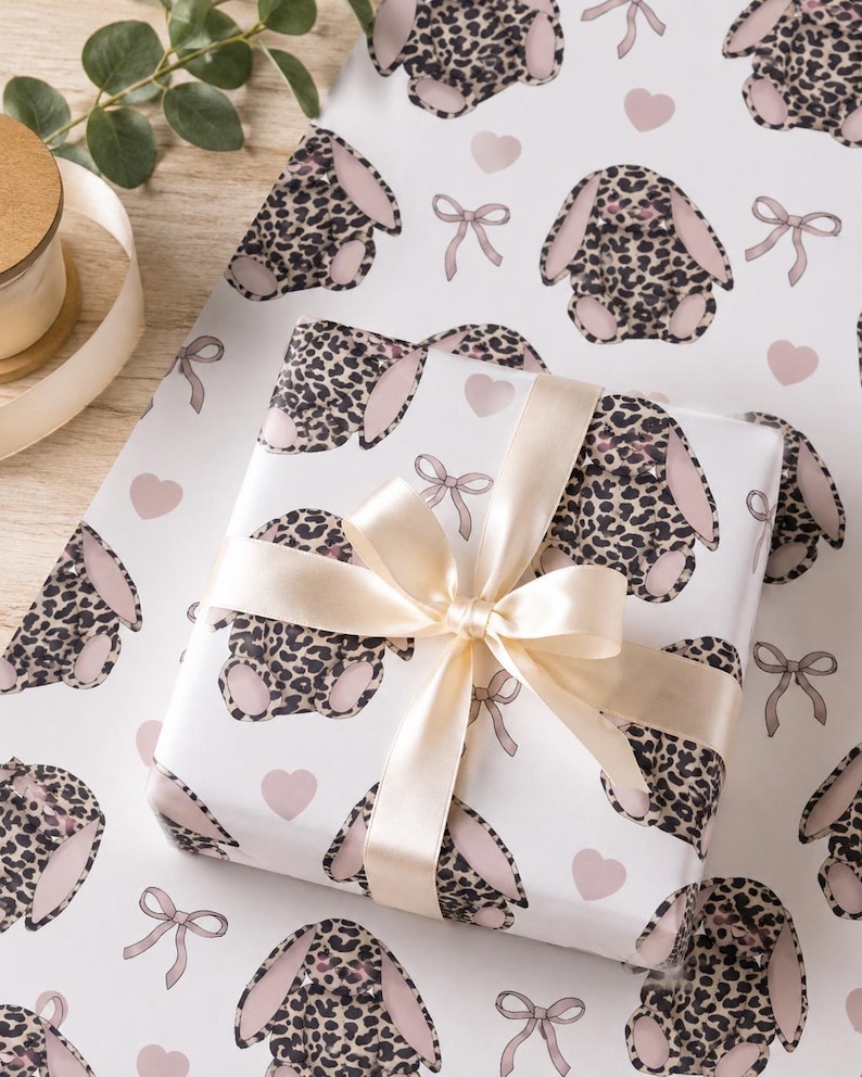 May include: White gift wrap featuring a repeating pattern of leopard print bunny rabbits, pink hearts, and bows. A wrapped gift is tied with a cream-colored satin ribbon. The overall theme is Easter or baby shower.