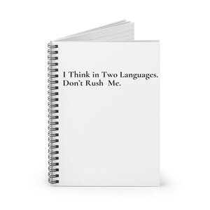 May include: A white spiral-bound notebook with the text "I Think in Two Languages. Don't Rush Me." printed in black. The notebook is open, showing blank white pages.