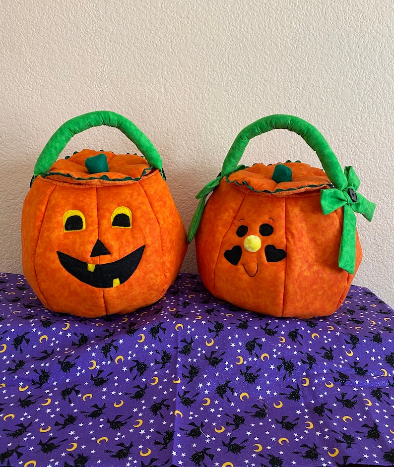 May include: Two fabric pumpkin-shaped baskets with green handles. The left basket features a black jack-o'-lantern face with yellow eyes and a tooth. The right basket has a face with heart-shaped eyes, a yellow nose, and a green side bow.