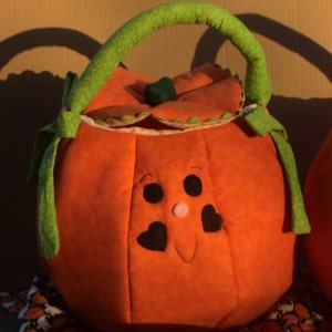 May include: A round, orange fabric pumpkin basket featuring a green handle and a lid with a small green stem. The pumpkin has a stitched face with two black circular eyes, two black heart-shaped cheeks, and a small pink nose on the front panel.
