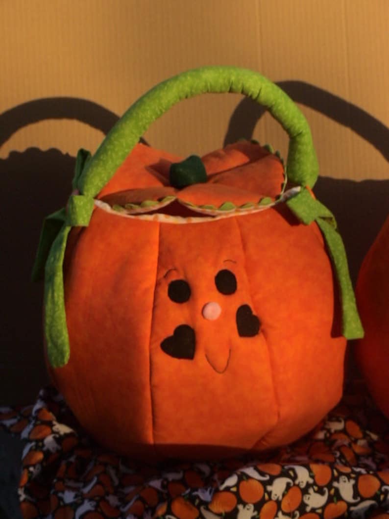May include: A round, orange fabric pumpkin basket featuring a green handle and a lid with a small green stem. The pumpkin has a stitched face with two black circular eyes, two black heart-shaped cheeks, and a small pink nose on the front panel.
