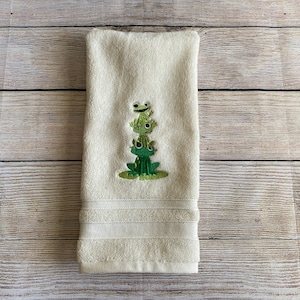 May include: A white hand towel with an embroidered design of three green frogs stacked on top of each other.
