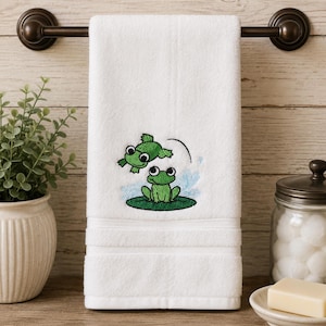 May include: A white hand towel featuring an embroidered design of two green frogs. One frog sits on a dark green lily pad while the other frog is depicted mid-air above it, with a curved black line indicating a jumping motion and blue water splashes.
