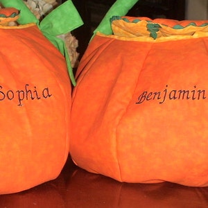 May include: Two orange fabric pumpkin-shaped bags with green handles and ruffled tops. Each bag features black embroidered text, with one displaying the name Sophia and the other displaying the name Benjamin on the front surface.