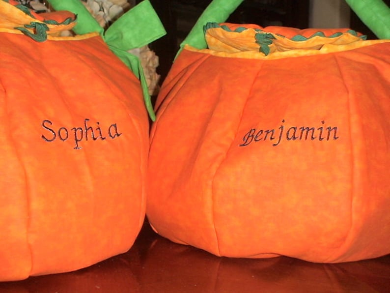 May include: Two orange fabric pumpkin-shaped bags with green handles and ruffled tops. Each bag features black embroidered text, with one displaying the name Sophia and the other displaying the name Benjamin on the front surface.