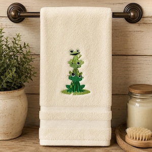 May include: A cream-colored hand towel featuring an embroidered design of three stacked green frogs sitting on a lily pad. The towel has a textured border near the bottom edge and is hanging from a dark metal rod.