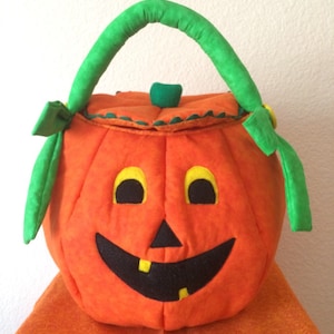 May include: A soft, orange fabric pumpkin bucket with a green handle and a lid. The pumpkin features a black felt face with yellow eyes and two yellow teeth in a wide, smiling mouth. The top of the lid has a small green stem accent.