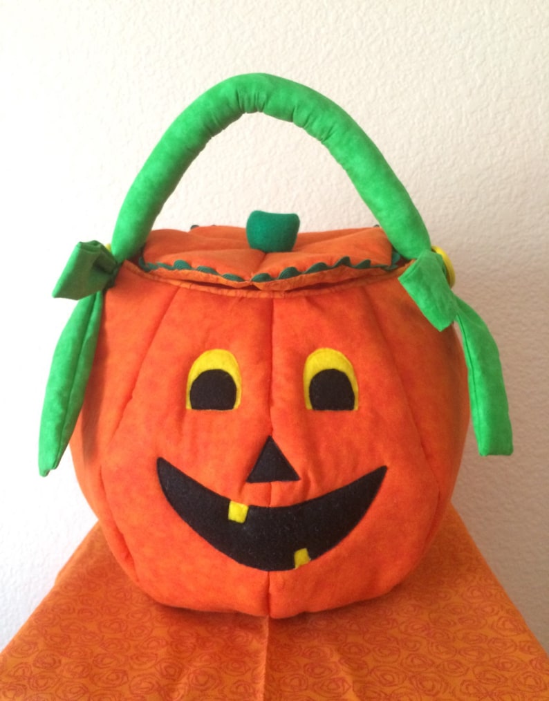 May include: A soft, orange fabric pumpkin bucket with a green handle and a lid. The pumpkin features a black felt face with yellow eyes and two yellow teeth in a wide, smiling mouth. The top of the lid has a small green stem accent.
