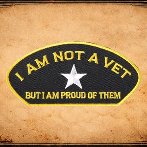 May include: A black patch with yellow trim and text that reads "I AM NOT A VET BUT I AM PROUD OF THEM." A white star is in the center of the patch. The patch is set against a brown background with a decorative border.