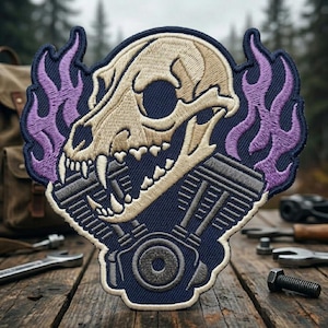May include: An embroidered patch featuring a beige animal skull positioned above a grey mechanical engine component, all set against a dark blue background with purple flames rising on both sides of the skull and a white border outline.