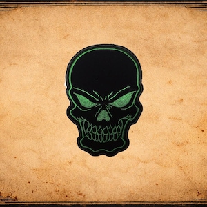May include: Black skull patch with green embroidered details. The skull has a menacing expression with detailed eye sockets and teeth. The patch is centered on a textured, tan background with a decorative border.