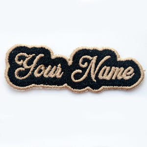 May include: A black embroidered patch with the words "Your Name" in a cursive font. The text is in a tan color and is outlined with a tan border. The patch is on a white background.