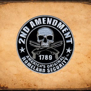 May include: A circular patch with the text "2ND AMENDMENT" in white on a black background. The patch features a skull, crossed pistols, the year "1789", and the words "AMERICA'S ORIGINAL HOMELAND SECURITY". The background is a distressed, aged paper.