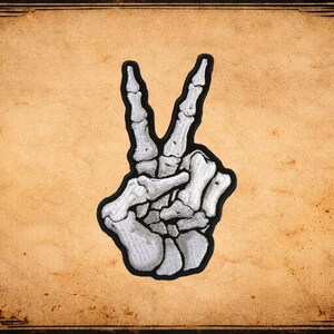 May include: A patch featuring a white, skeletal hand making a peace sign. The hand is detailed with bone structures and outlined in black. The patch is set against a vintage, tan background with a decorative border.