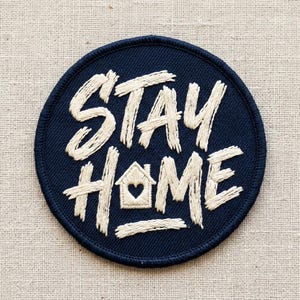 May include: A navy blue embroidered patch with the words "STAY HOME" in cream-colored, textured lettering. The word "HOME" features a house icon with a heart. The patch is circular and has a stitched border.