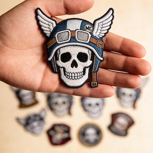 May include: Embroidered patch featuring a skull wearing a blue and white motorcycle helmet with goggles and wings. The patch has brown and white accents and the letters "MC". Other patches with skull designs are visible in the background.