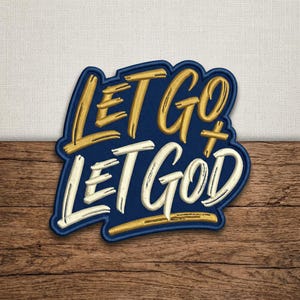 May include: Embroidered patch with the words "LET GO LET GOD" in gold and white script. The patch has a navy blue background with a gold border and is set against a wooden surface.