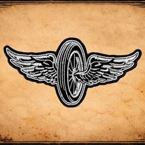 May include: Embroidered patch featuring a black and white winged wheel design. The patch has a detailed, textured appearance with the wings extending outward from the wheel. The background is a textured, aged paper.
