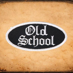 May include: An oval-shaped black patch with the words "Old School" in white gothic lettering. The patch has a white stitched border. The background is a textured, sepia-toned paper with decorative corner elements.