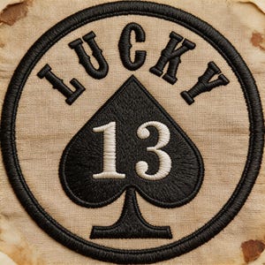 May include: Embroidered patch with the words "LUCKY" and "13" in black thread. The number 13 is inside a black spade shape. The patch is circular with a black border and is on a tan fabric background.