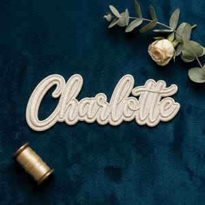 May include: Cream-colored embroidered patch with the name "Charlotte" in a cursive script. The patch is on a dark teal velvet surface, with a spool of gold thread and a rose with greenery in the background.
