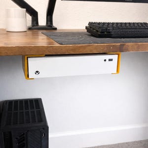 May include: A white gaming console is mounted under a wooden desk with a yellow bracket. A black keyboard, mousepad, and monitor are on the desk. A black computer case sits below the desk.