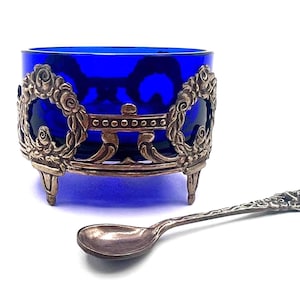 May include: A deep cobalt blue glass bowl encased in an ornate silver-toned metal holder featuring floral wreath motifs and three small feet, accompanied by a matching silver-toned metal spoon with a detailed floral handle design.