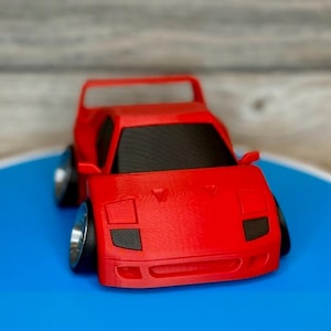 May include: A red toy car with black windows and silver wheels. The miniature vehicle has a spoiler and a low profile, with a detailed front end. The car is set against a blue and gray background.