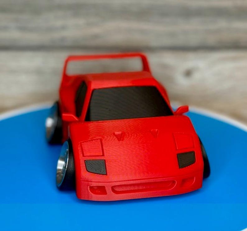 May include: A red toy car with black windows and silver wheels. The miniature vehicle has a spoiler and a low profile, with a detailed front end. The car is set against a blue and gray background.