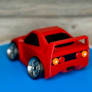 May include: A small, red toy car with silver wheels and a rear spoiler. The car has black windows and orange and red taillights. The toy car is on a blue surface.