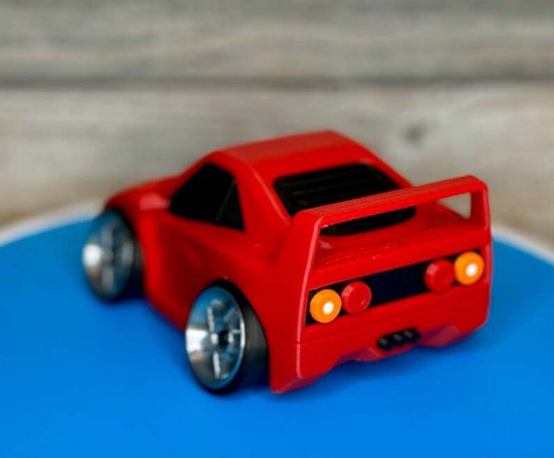 May include: A small, red toy car with silver wheels and a rear spoiler. The car has black windows and orange and red taillights. The toy car is on a blue surface.