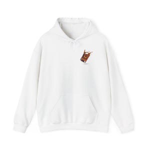 May include: A white hooded sweatshirt featuring a small graphic on the upper left chest. The graphic depicts a tilted glass filled with a dark brown liquid, showing a dynamic splash effect with amber and black tones against the plain white fabric.