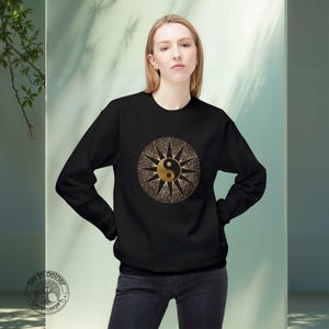 Könnte beinhalten: Schwarzes Sweatshirt mit einem goldenen Yin-Yang-Symbol in einem Sonnenmuster. Die Grafik ist auf der Brust zentriert. Das Sweatshirt hat lange Ärmel und ist aus weichem Material. Das Model trägt eine dunkle Jeans.
