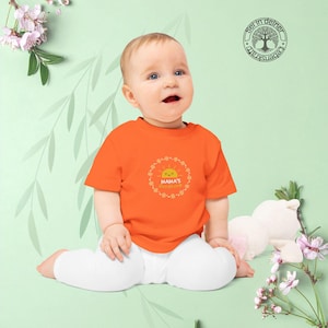 Könnte beinhalten: Ein orangefarbenes Baby-T-Shirt mit dem Text "Mama's Sunshine" in einem kreisförmigen Design mit einer lächelnden Sonne. Das Baby trägt eine weiße Hose und sitzt vor einem hellgrünen Hintergrund mit floralen Akzenten.
