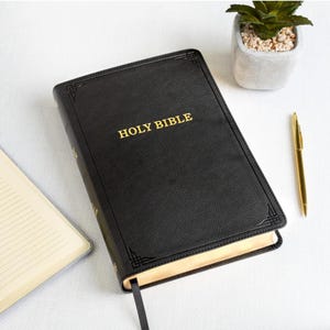 Puede incluir: Una Santa Biblia negra encuadernada en cuero con letras doradas y un marcador a juego. El libro está sobre una superficie blanca junto a un bolígrafo dorado y una pequeña planta en maceta.