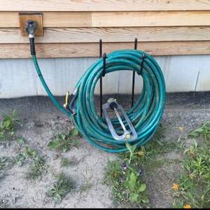 May include: A green garden hose is coiled on a metal hose holder. The hose is attached to a water spigot on a wooden wall.