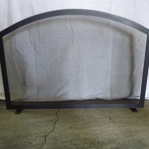 Puede incluir: Pantalla de chimenea de metal negro con parte superior arqueada y diseño de malla. La pantalla está de pie sobre un suelo de hormigón.