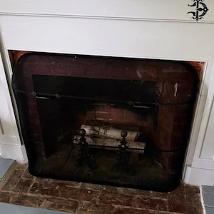 Puede incluir: Una chimenea con una pantalla de malla negra, interior de ladrillo y una repisa blanca. La chimenea contiene troncos y detalles metálicos decorativos. La repisa tiene un emblema negro decorativo en el lado derecho.