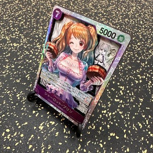 Pode incluir: Uma carta colecionável retangular com um personagem de estilo anime com cabelo castanho claro em marias-chiquinhas, vestindo um vestido roxo claro e segurando um pequeno bolo. A carta exibe o número 7, o valor 5000 e texto em japonês.