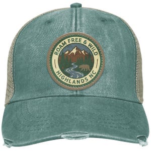 May include: A distressed teal trucker hat with a mesh back and a curved brim. The front features a circular patch with the text "ROAM FREE & WILD HIGHLANDS NC" around a mountain and bear graphic.