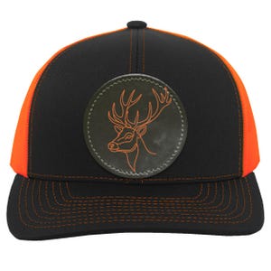 May include: Black and orange trucker hat with a deer head patch. The hat features a black front panel, an orange mesh back, and a curved brim. The patch is a circular leather design with an orange deer outline.